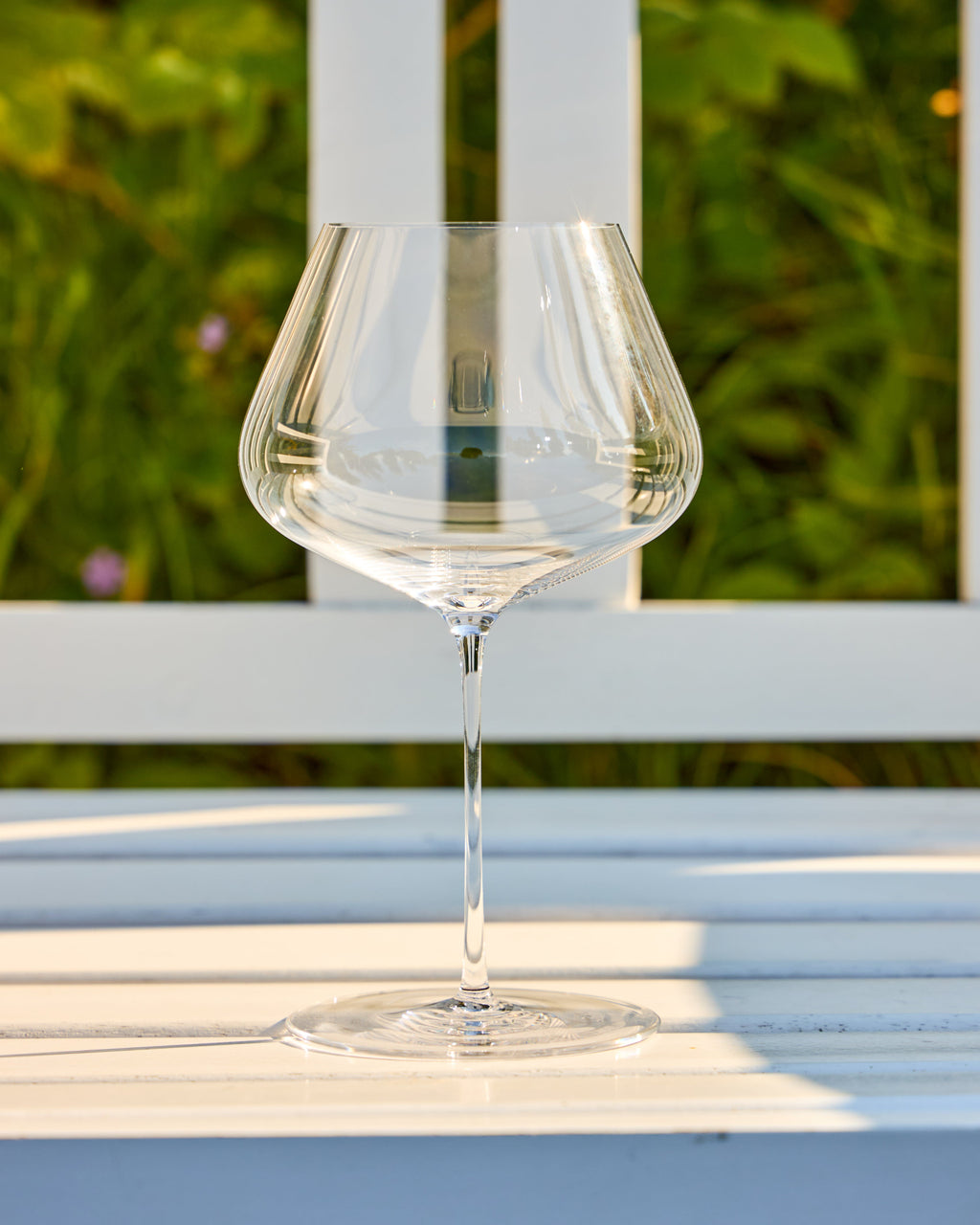 A clear stemmed Zalto Balance wine glass with a wide, rounded bowl sits on a white wooden table outdoors, with a blurred green garden and white fence in the background.