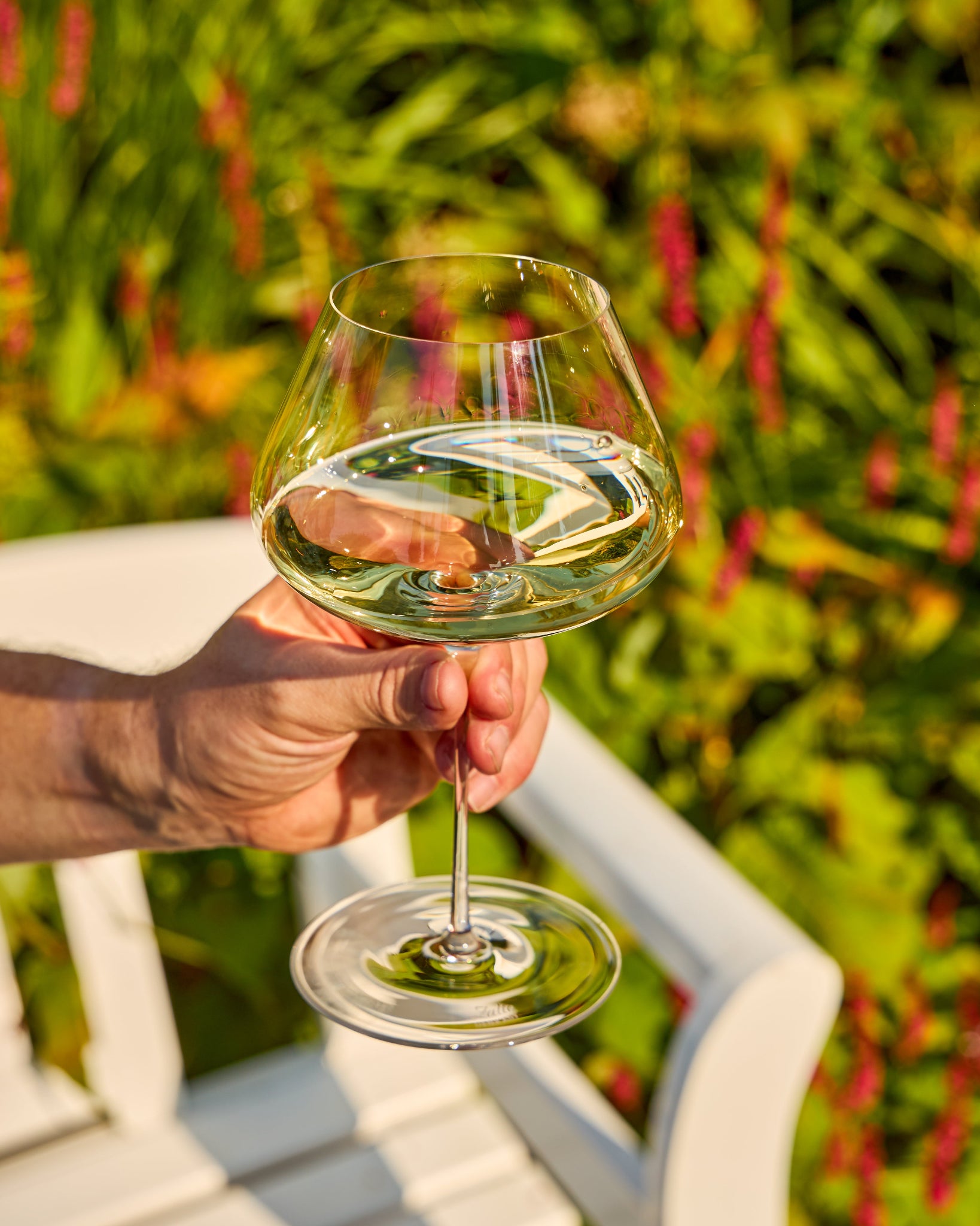 A hand holds a clear stemmed Zalto Balance wine glass with a wide bowl, filled with a pale green liquid, in front of a white chair and a blurred garden background with green foliage and pink flowers.